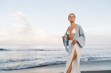 Muhteşem beyaz kadın plajda sakin ve kaygısız bir şekilde yürürken tatilin tadını çıkarıyor. Beyaz yaz giysileri içinde güzel, sağlıklı bir kadın tatil boyunca deniz kıyısında gezer.