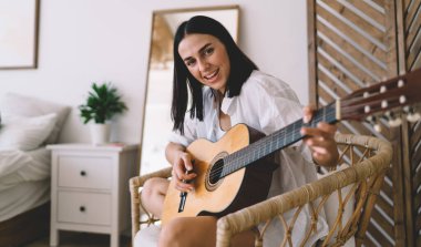 Gülümseyen ve gitar çalmayı öğrenen esmer, çekici bir kadın ev içinde rahat bir şekilde oturuyor. Genç mutlu kadın müzik aleti performansıyla hobisinin keyfini çıkarıyor hasır sandalyede dinleniyor.