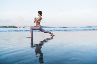 Modayı takip eden sportif giyinen sakin bir kadın. Kıyı şeridinde yoga eğitimi alırken dinleniyor. Uyum ve dengeyi hissediyor.