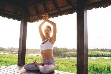 Spor kıyafetleri içinde beyaz bir kadın lotus pozisyonunda meditasyon yapıyor ve Hatha yoga uygulamasını düşünüyor. Çekici, sağlıklı bir kız gün ışığında huzurlu aydınlanmanın tadını çıkarıyor.