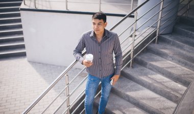 Ofis binasının yanındaki merdivenlerde cep telefonu ve kahve taşıyan Asyalı adam. Dinlenme ve işten ayrılma kavramı. Modern, başarılı bir adamın fikri. Genç bir erkek başka tarafa bakıyor. Güneşli bir gün