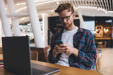 Genç beyaz adam masada cep telefonundan bir şey izliyor. Çevrimiçi eğitim ve iş kavramı. Modern, başarılı bir adamın fikri. Gözlüklü adam geniş ofiste çalışıyor ya da çalışıyor.