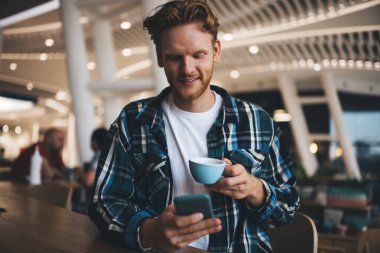 Beyaz adam akıllı telefondan izliyor ve bulanık ofiste fincandan çay ya da kahve içiyor. Dinlenme ve işten ayrılma kavramı. Modern, başarılı bir adamın fikri. Masada oturan gülümseyen genç erkek.
