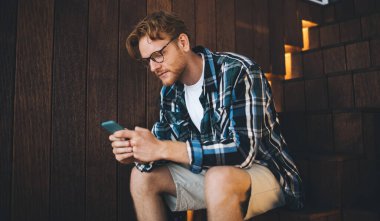 Genç, beyaz bir adam merdivenlerde oturmuş iş yerinde akıllı telefonları izliyor. Uzaktan eğitim ve iş kavramı. Modern, başarılı bir adamın fikri. Gözlüklü, ciddi sakallı bir erkek.