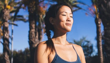 Palmiyelerle sokakta nefes almak için Pranayama yaparken gözlerini kapayan Asyalı kadın, sakin kadın yogi hissi bütünsel iyileşme ve geri çekilme pratiği sırasında aydınlanma hissi.