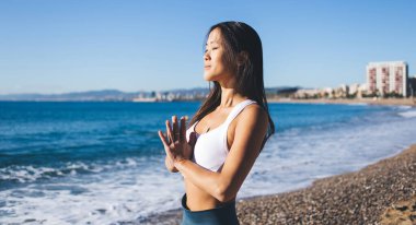 Sakin Asyalı kadın namaste 'de dua etmeye devam ediyor. Deniz kıyısında meditasyon yaparken güneş enerjisi alıyor. Beyaz bluzlu genç kadın yoga yaparken Zen ve farkındalık hissediyor.