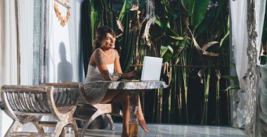 Otelin terasında dizüstü bilgisayarda yazı yazan ve izleyen beyaz bir kadın. Serbest çalışma ve uzak çalışma kavramı. Tahta masada oturan dövmeli güzel bir kız. Güneşli bir gün. Bali adası