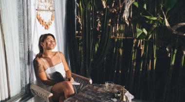 Not defteriyle hayalperest genç kadın tatil köyünün terasında güneşli bir günün tadını çıkarıyor. Turizm, tatil ve hafta sonu. Çekici gülümseyen beyaz kız. Bali adasında huzurlu ve huzurlu bir yaşam tarzı