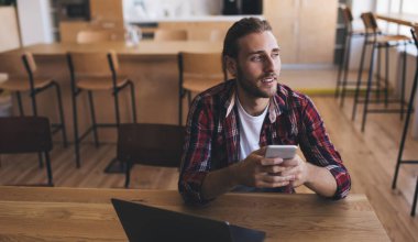 Modern dizüstü bilgisayarı ve akıllı telefon aleti olan düşünceli bir erkek programcı. Düşünceli bir şekilde başka yere bakıyor ve grafik tasarımı için web içeriğini düşünüyor.