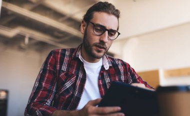 Üniversite kampüsünde öğrenim görürken eğitim bilgilerini analiz eden beyaz bir öğrenci, görsel koruma için optik gözlüklü zeki bir erkek, organizasyonu planlarken notlar okuyor.