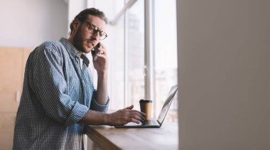 Görme koruması için optik gözlüklü, kafası karışmış beyaz adam cep telefonu teknolojisini kullanarak dizüstü bilgisayar sorunlarını tartışıyor, yetenekli yazılım geliştiricisi akıllı telefondan iş konuşması yapıyor.