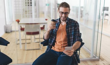 Milenyum Kafkas işadamı akıllı telefondan kulaklıkla müzik dinliyor ve seçiyor. Modern başarılı adam kavramı. Gözlüklü, gülümseyen genç girişimci ofiste oturuyor ve çalışıyor.