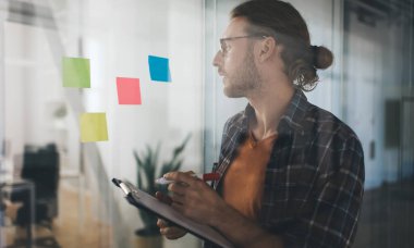 Milenyum Kafkas işadamı, iş sırasında yapışkan kağıda bakıp panoya yazı yazıyor. Modern başarılı adam kavramı. Açık uzay ofisinde gözlüklü genç bir girişimci.