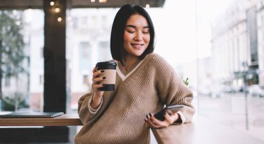 Akıllı telefon kullanıp kafede kahve içen Asyalı bir kız. Genç gülümseyen milenyum esmer kadın pencerenin yanındaki masada duruyor. Dinlenmek, boş zaman ve boş zaman kavramı. Modern kadın yaşam tarzı