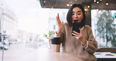 Akıllı telefondan izleyen ve kafede el kol hareketi yapan Asyalı kız şok oldu. Genç milenyum esmer kadın pencerenin yanındaki masada duruyor. Dinlenmek, boş zaman ve boş zaman kavramı. Modern kadın yaşam tarzı