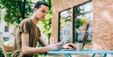 Sokak kafeteryasındaki masada oturmuş laptopları karıştırıyor ve cep telefonundan mesajlar yolluyor.