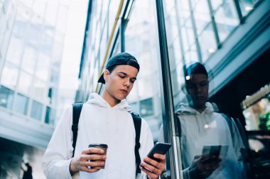 Düşük açılı, sıradan kıyafetli, sırt çantalı, elinde kağıt bardak sıcak içecek ve cep telefonuyla sokakta yürüyen genç bir adam.