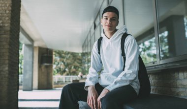 Beyaz kapüşonlu beysbol şapkalı, sırt çantalı, terasta oturan ve kameraya bakan genç pozitif bir adam.