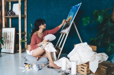Çekici genç bayan ressam resim çiziyor ve hobisinin rahatlatıcı sürecinden zevk alıyor. Yaratıcı ve yetenekli esmer kadın stüdyoda resim yapıyor. Petrolle çizim yapmayı öğrenmek
