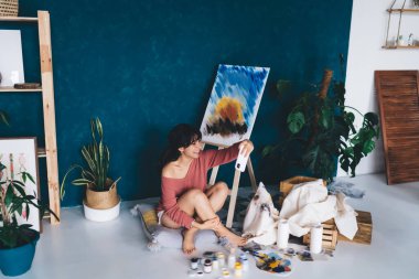 Tasasız genç bayan sanatçı resim sehpası yakınında akıllı telefon kamerası için poz veriyor ve blogunda hobisini gösteren resimler yapıyor. Yaratıcı gülümseyen esmer kadın, yağlı boya resminin yanında kendini fotoğraflıyor. 