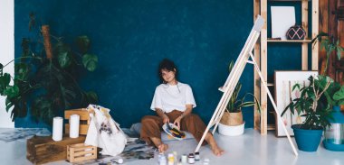 Çekici genç bayan sanatçı stüdyo zemininde oturmuş hobisinin keyfini çıkartıyor. Yaratıcı ve yetenekli esmer kadın sehpada resim yaratıyor. Petrolle çizim yapmayı öğrenmek