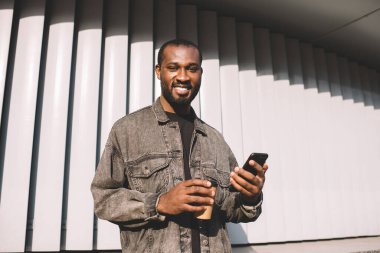 Modern cep telefonu teknolojisiyle neşeli erkek blog yazarının yarım boy portresi ve şehir merkezinde boş zamanlarında kameraya gülümseyen bir fincan kafein içeceği, mutlu koyu tenli kullanıcı.