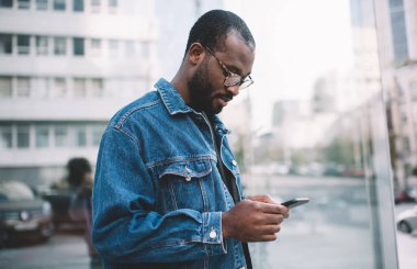 Sosyal medyadaki içerik yayınlarını kontrol etmek için cep telefonu uygulamasını kullanan koyu renk tenli genç turist, akıllı telefon aracılığıyla yeni nesil Afro-Amerikan hipster adam ağı