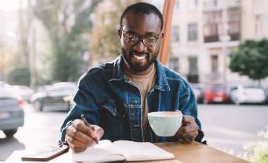 Gündüz vakti kaldırım kafesinde fikir ürettiği için gülümseyen koyu tenli mutlu erkek yazar. Optik gözlüklü neşeli hippi adam elinde kahve fincanı tutarken göz düzeltmesi yapıyor.