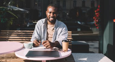 Bluetooth kulaklıklı neşeli erkek öğrenci, eğitim kitaplarını dinlerken ödev testi yapıyor. Kulaklık takmış, kulaklık takmış mutlu koyu tenli hippi adamın resmi.