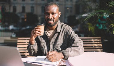 Elektronik kulaklıklar takmış neşeli koyu tenli bir öğrenci uzaktan kumandayla ve kitapla mutlu hipster bir adamın kahve saatinde Muisc 'in keyfini çıkarırken ses kayıtlarını dinliyor ve kameraya gülümsüyor.