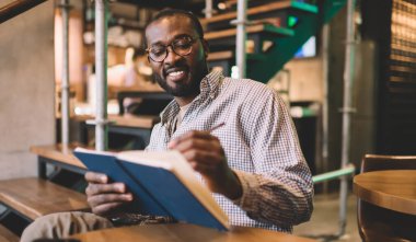 Neşeli koyu tenli erkek eğitim bilgisi yazarken üniversite ödevlerini ders kitabında yapıyor. Görme engeli için optik gözlüklü mutlu Afrikalı Amerikalı öğrenci içeride notlar alıyor.