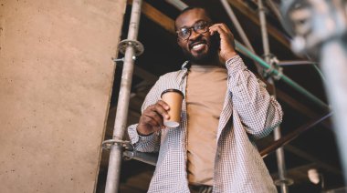 Optik gözlüklü siyahi hippi adamın yarım boy portresi. Dostça cep telefonuyla konuşurken kameraya gülümsüyor. Kahve saatinde Afrika kökenli Amerikalı bir adam cep telefonuyla bağlantı kuruyor.