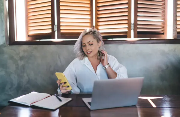 stock image Millennial blonde woman browsing text content on modern smartphone gadget using 4g wireless in coworking space, young female blogger with laptop and notepad using cellular device for online booking