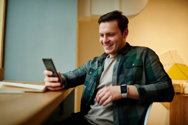 Genç gülümseyen erkek, kafenin içinde otururken akıllı telefondan mesaj okuyor. Kafkasyalı adam cep telefonu uygulaması kullanıyor 