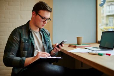 Akıllı adam akıllı telefon uygulamasındaki elektronik versiyonu düzeltirken belgeleri okuyor. Gazetelerle çalışan ve kahve dükkanında teknoloji kullanan serbest çalışan bir erkek.