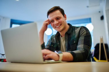 Genç gülümseyen adam internette çalışırken dizüstü bilgisayarda oturuyor. Erkek öğrenci netbook ile öğreniyor. Web sitesinde makale okunuyor