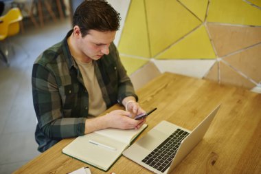 Genç düşünceli bir adam dizüstü bilgisayar ve not defteriyle masada otururken cep telefonuna mesaj atıyor. Serbest çalışmak, teknolojiyi kullanmak. Akıllı telefon uygulaması