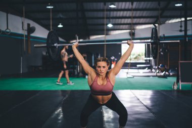 Spor stüdyosunda vücut geliştirme egzersizi yaparken halter kaldıran güçlü bir kadının portresi, ağırlık kaldırırken ve egzersiz yaparken kameraya bakan beyaz eşofmanlı bir bayan atlet.