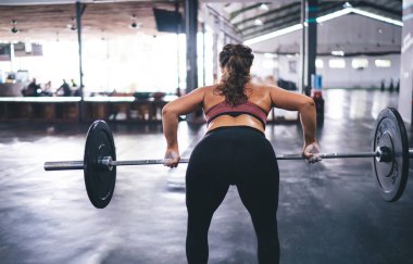 Spor stüdyosunda vücut geliştirme egzersizi yaparken halter kaldıran formda bir kadının arka planda duruşu. Eşofmanlı kadın atlet, ağırlık kaldırma egzersizi ve ağırlık kaldırma egzersizi yapıyor.