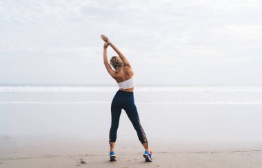 Uzunluk açısı geniş, spor vücutlu beyaz kadın açık havada antrenman yaparken deniz kıyısında esneme egzersizleri yapıyor. Genç bir kadın doğada spor yapıyor ve sağlıklı yaşam tarzının tadını çıkarıyor. Sahilde ısın.