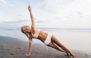 Uzunluk açısı geniş, spor vücutlu beyaz kadın açık havada antrenman yaparken deniz kıyısında esneme egzersizleri yapıyor. Genç bir kadın doğada spor yapıyor ve sağlıklı yaşam tarzının tadını çıkarıyor. Sahilde ısın.