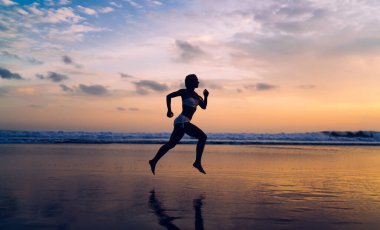 Uzunluk açısı, açık havada antrenman yaparken deniz kıyısında koşan atletik vücutlu bir kadın silueti. Genç bir kadın doğada spor yapıyor ve sağlıklı yaşam tarzının tadını çıkarıyor. Koşucu gün batımında sahilde hareket eder.