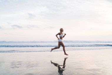 Tam uzunlukta, açık havada antrenman yaparken deniz kıyısında koşan beyaz tenli bir kadın. Genç bir kadın doğada spor yapıyor ve sağlıklı yaşam tarzının tadını çıkarıyor. Kaçak okyanus sahilinde hareket ediyor