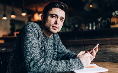 Kafe 'de eğitim görürken kameraya bakan modern hücresel aygıtlarla genç beyaz öğrencinin portresi. Mobil cihaz ve eğitim notlarıyla binlerce yıllık blogcu masada poz veriyor.