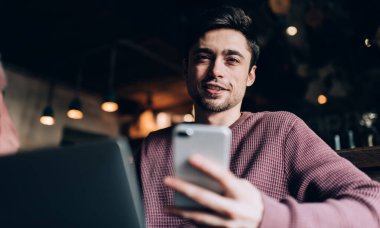 Modern akıllı telefon ve laptop teknolojisiyle yakışıklı beyaz blog yazarının portresi boş zamanlarında kameraya bakıyor, netbook ve mobil cihazlı genç hipster adam birlikte çalışma alanında poz veriyor.