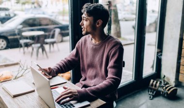 Kafe içinde modern dizüstü bilgisayar ve dijital cep telefonu teknolojisi kullanan, gündelik giyimli, düşünceli, beyaz tenli hippi bir adam. Netbook ve akıllı telefon düşünen düşünceli bir erkek programcı.