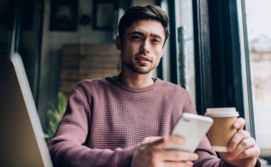 Modern dizüstü bilgisayarlı ve cep telefonlu yetenekli beyaz erkek programcı portresi netbook ve paket kupalı yetenekli hipster adam 4G 'ye bağlanmak için mobil uygulama kullanıyor.