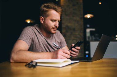 Kızıl saçlı genç adam web sitesinde çalışırken çizim defterine notlar yazıyor dizüstü bilgisayar ve akıllı telefon kullanıyor. Bin yıllık erkek öğrenci aletleri kullanarak öğreniyor. Serbest planlama projesi sorunları