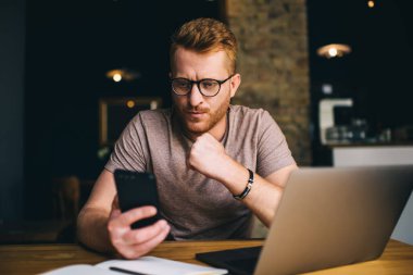 30 yaşında düşünceli bir kızıl adam bilgisayarını kapalı bir yerde açarken akıllı telefon uygulaması kullanıyor. İş yerinde serbest çalışma sırasında cep telefonundan okunan şaşkın erkek mesajı