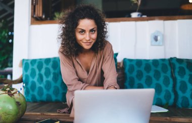 Gündüz vakti tropikal kafe terasında kameraya gülümseyen modern dizüstü bilgisayar teknolojisi olan Kafkas dijital göçebe portresi, netbook pozu veren bin yıllık kadın.